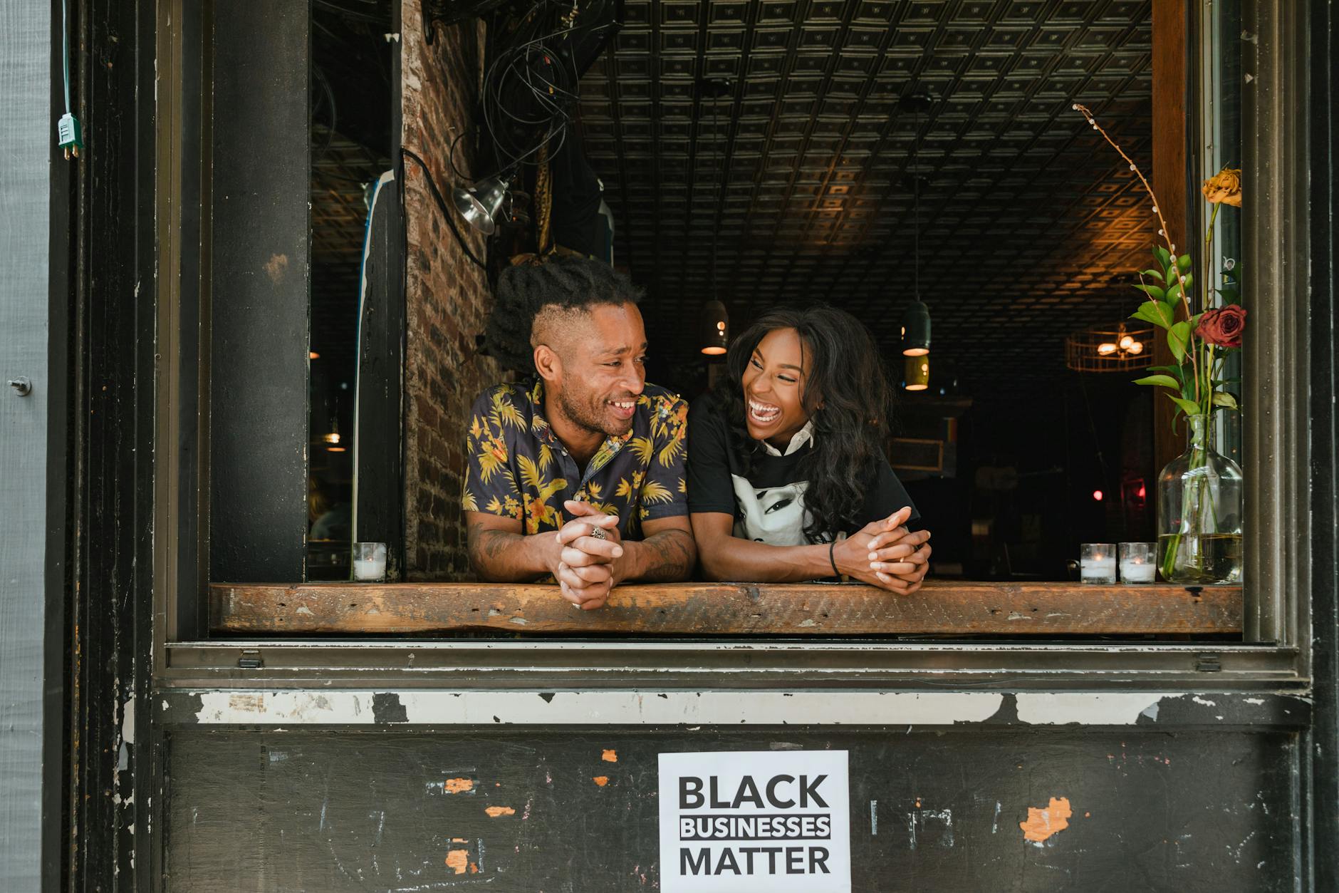 Two joyful business owners lean on a window sill with a 'Black Businesses Matter' sign visible.