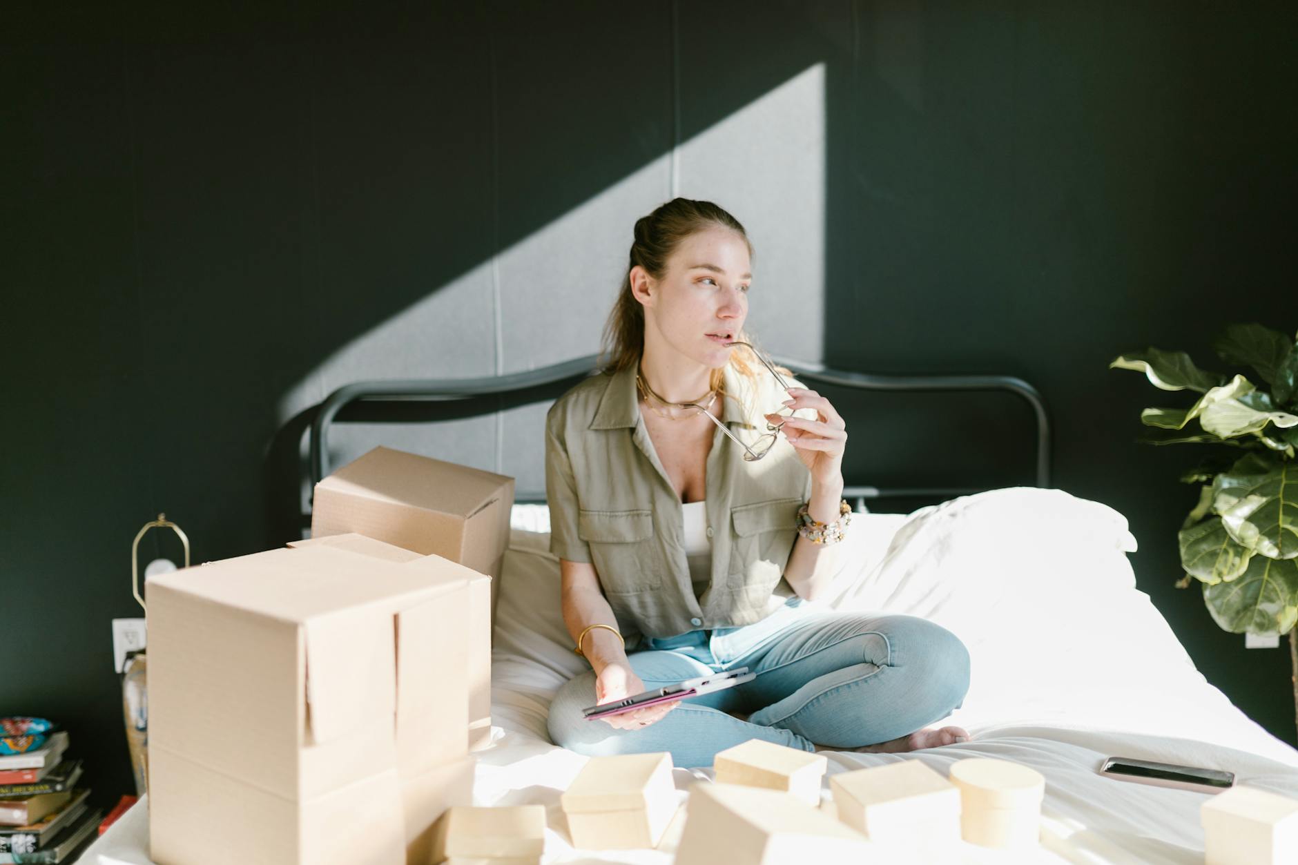 A woman sits with tablet and coffee, organizing her home-based e-commerce setup with boxes.