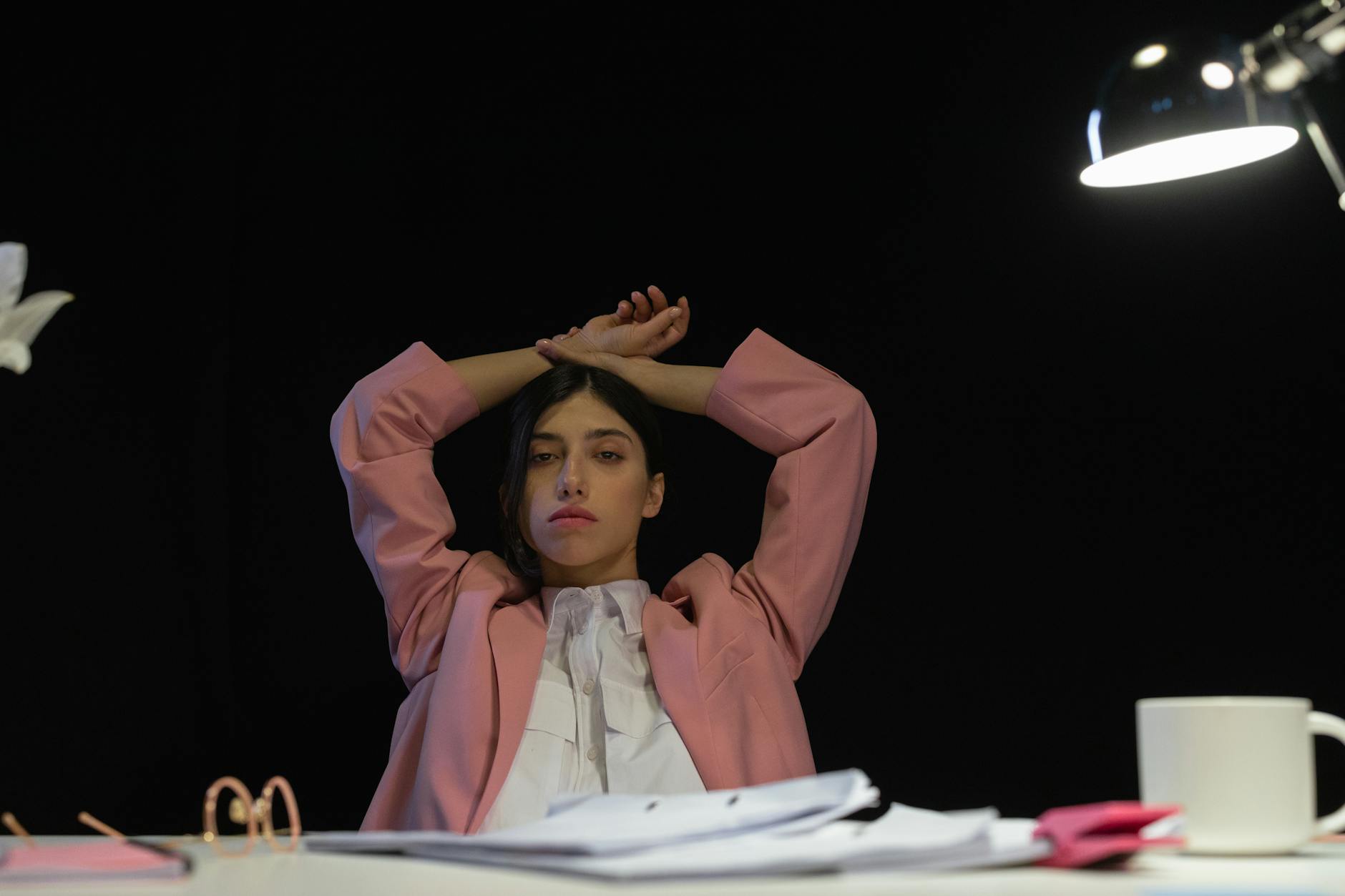 Woman in a pink suit sitting at a desk, arms raised, under a lamp, exuding confidence.