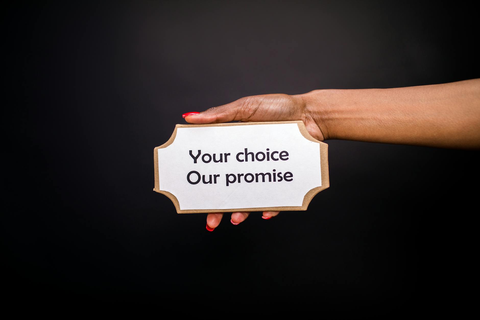A hand holds a sign saying 'Your choice, Our promise' against a dark background.