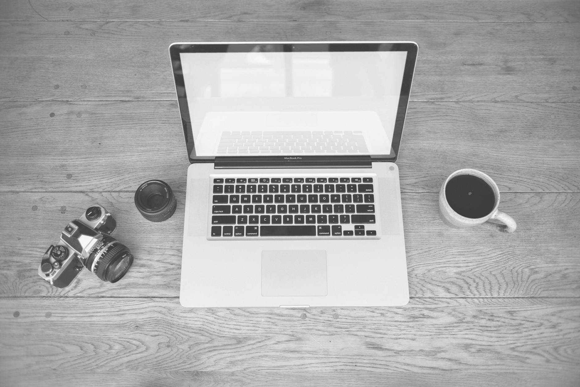 Monochrome image of a laptop, camera, lens, and coffee cup on a wooden desk