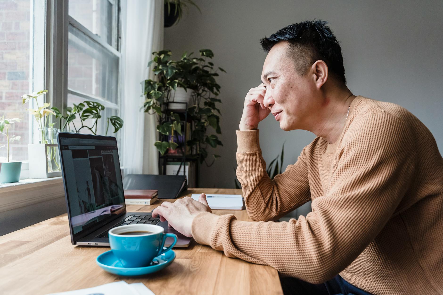 Asian man in casual attire working remotely on laptop at desk with coffee, focused and engaged.