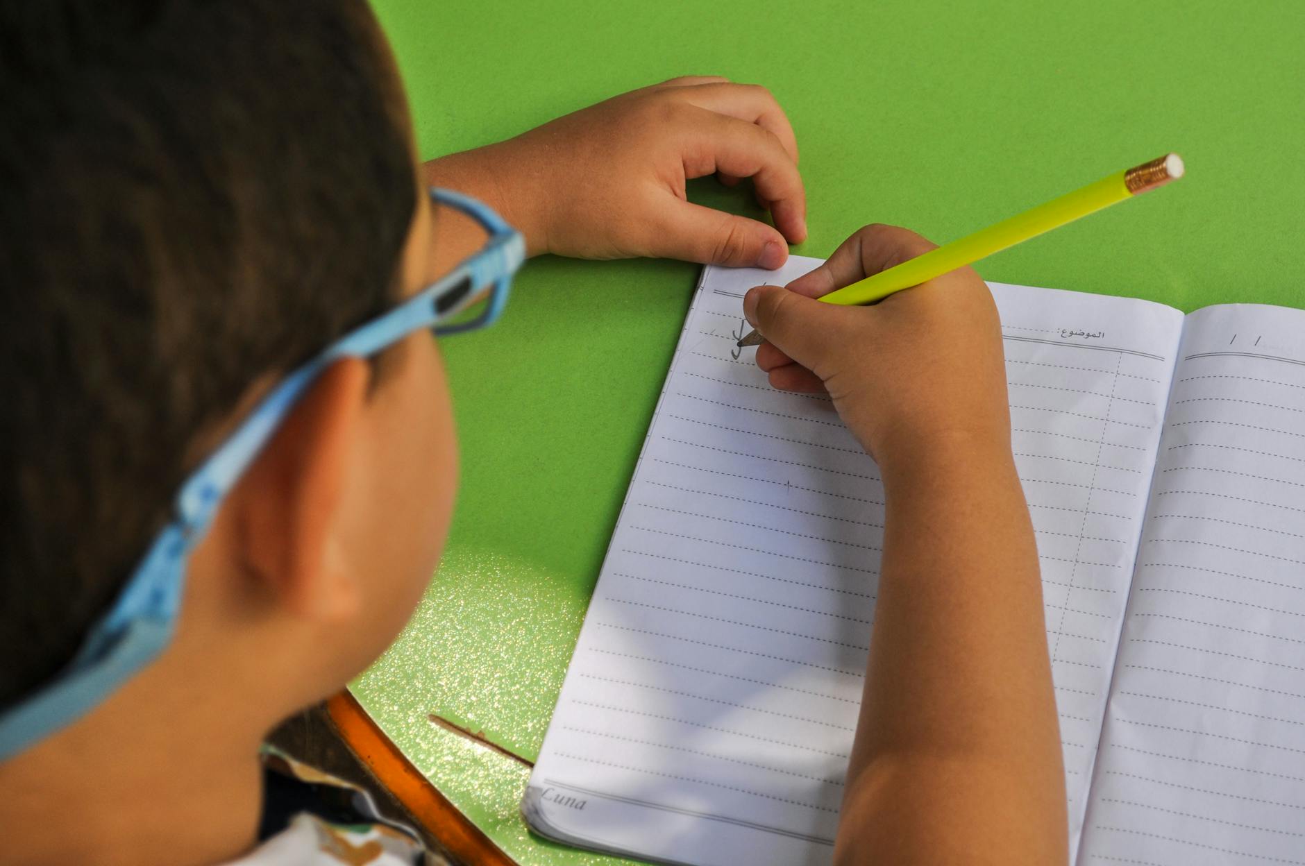 A child wearing glasses writes in a lined notebook with a yellow pencil on a green table.
