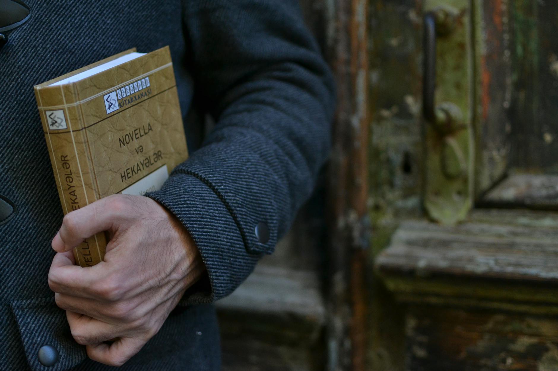A hand holding a book titled 'Novella və Hekayələr' in front of a weathered wooden door.