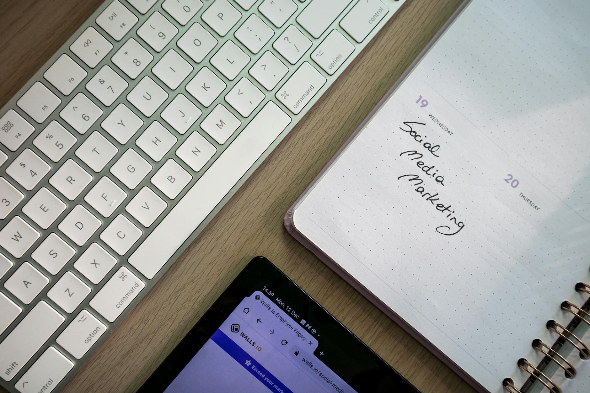 Top view of keyboard, planner with social media marketing note, and tablet on a desk.