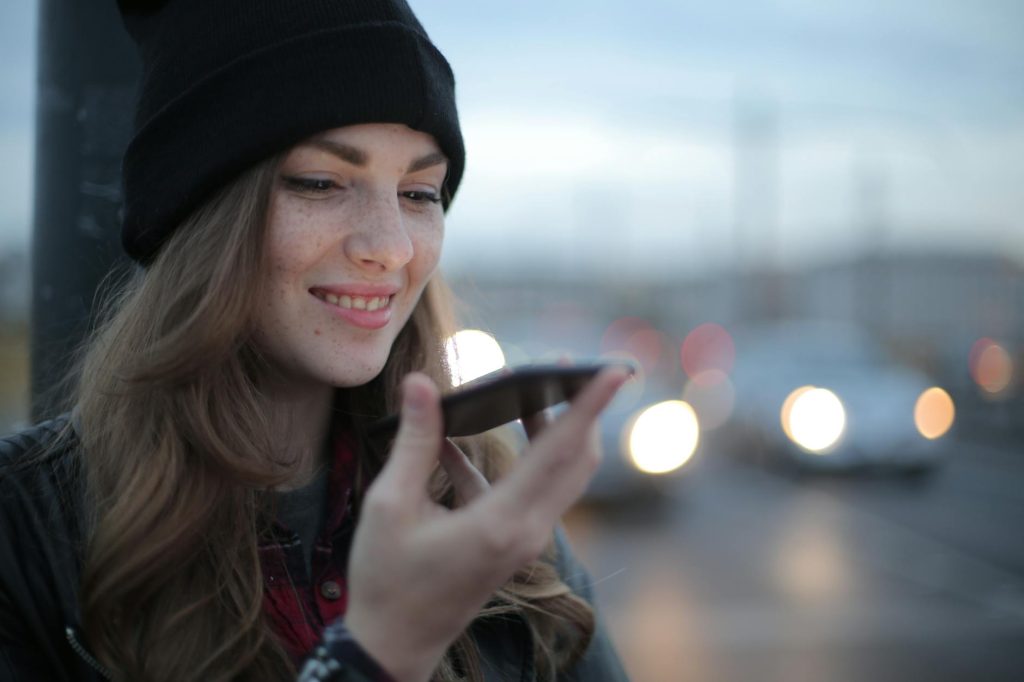 Crop millennial female in jacket and warm hat focusing on smartphone screen and smiling while making phone call against blurred cars driving along street road in evening city